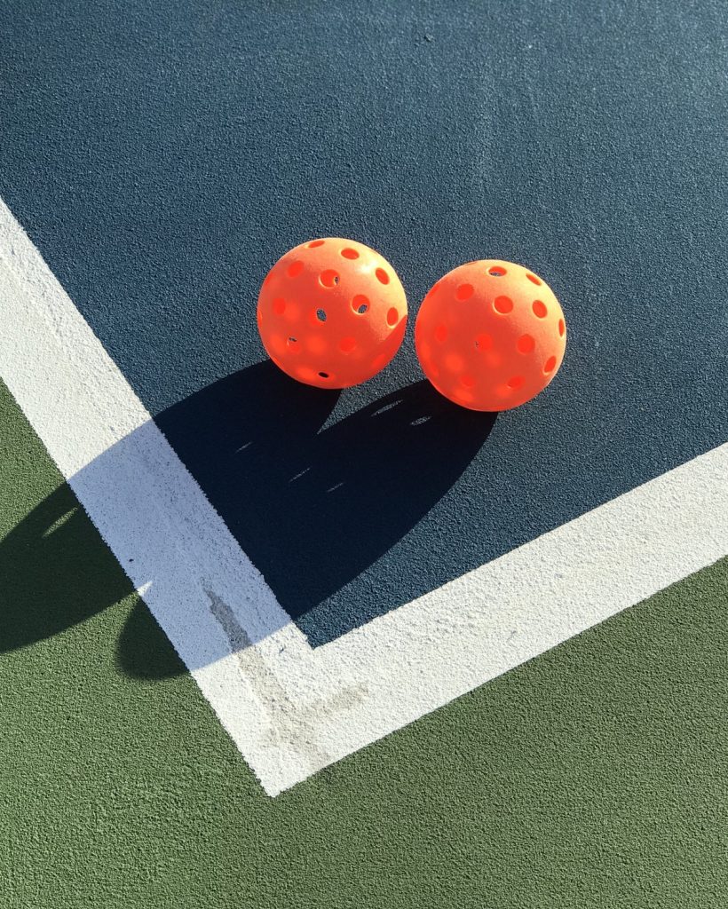 Nominated Two orange pickleballs in the corner of a pickleball court. sunlight forming shadows.